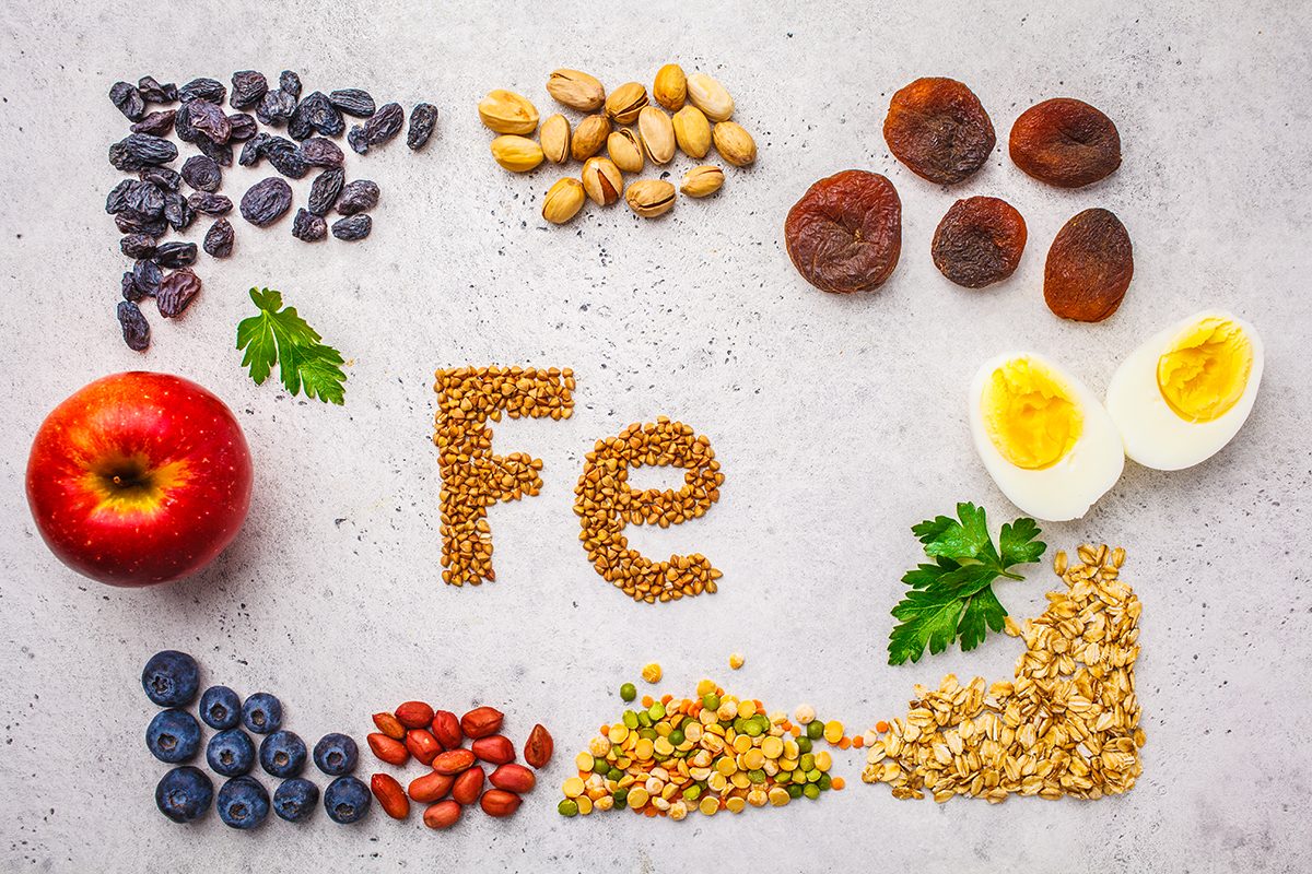 An assortment of iron-rich foods, including fruits, nuts, lentils and eggs, are displayed on a surface with buckwheat, that has been arranged to read "Fe", in the centre