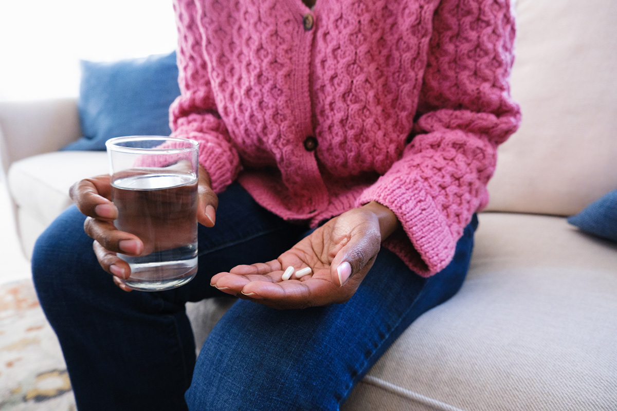 A woman is sitting on a sofa, holding a glass of water in one hand and two capsules in the other