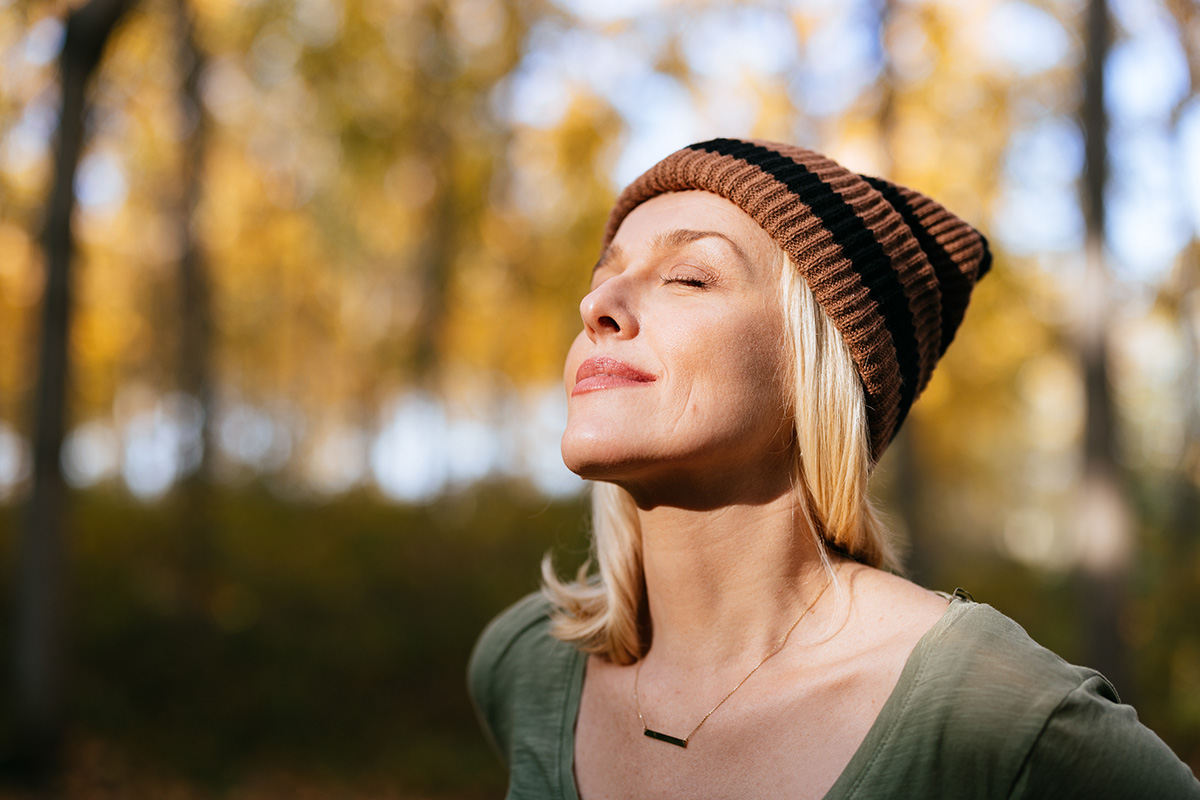 A woman is standing in an autumnal woods, taking a deep breath with her eyes closed