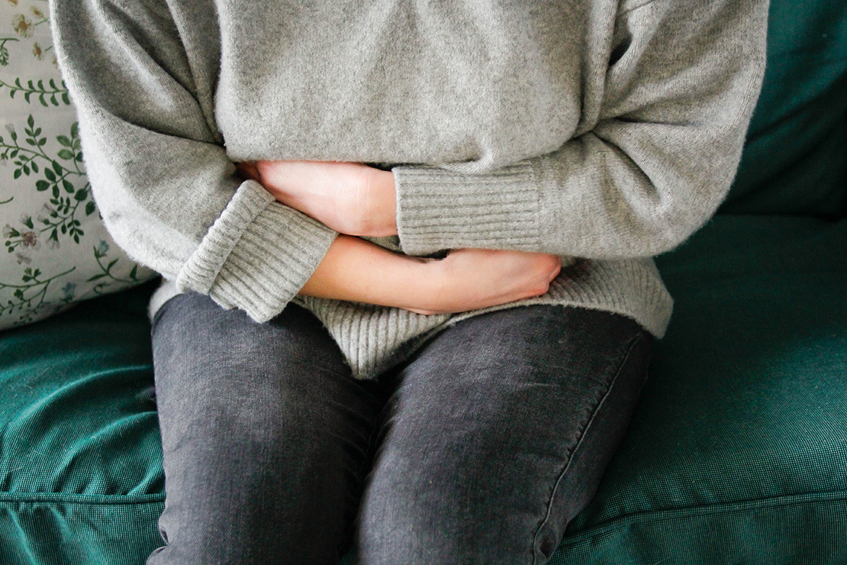 A woman is sitting on a sofa with her arms crossed over her stomach, suggesting stomach pain