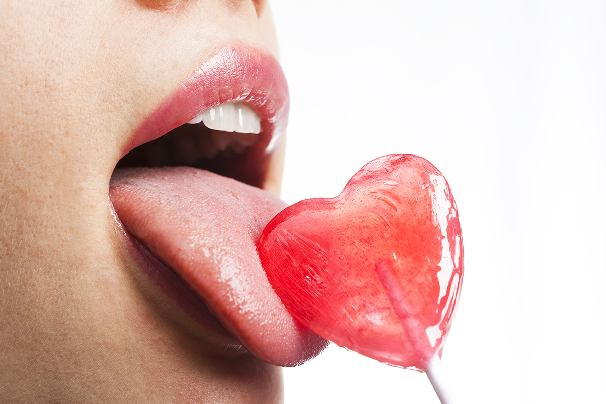 A close up of a woman's mouth licking a heart-shaped lollipop. 