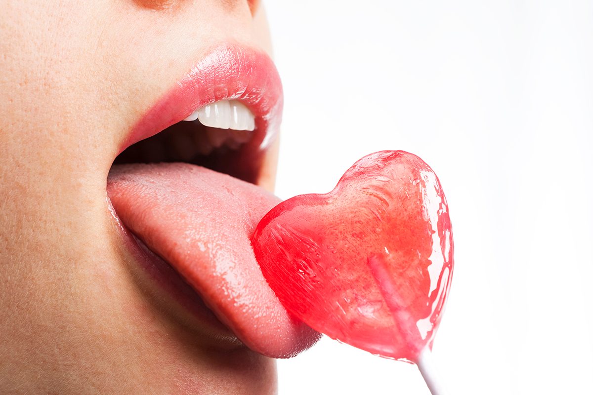 A close up of a woman's mouth licking a heart-shaped lollipop. 