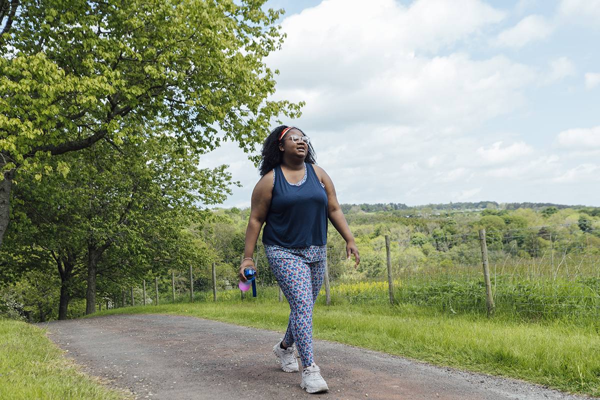 A woman in a blue vest top and patterned leggings walks along a scenic path, surrounded by trees and rolling hills.