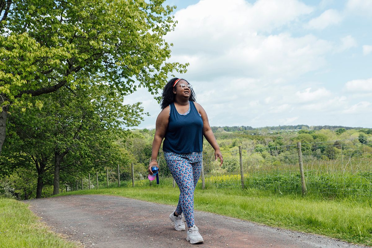 A woman in a blue vest top and patterned leggings walks along a scenic path, surrounded by trees and rolling hills.