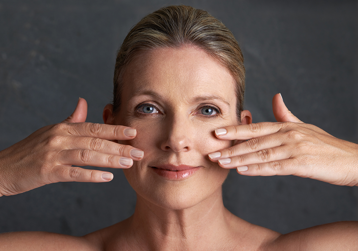 A middle-aged woman touching her face with both hands against a dark background