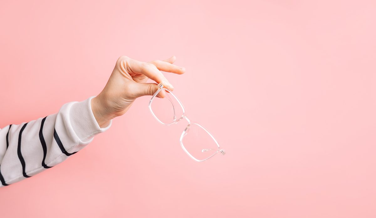 A hand holds glasses with a clear frame against a pink background