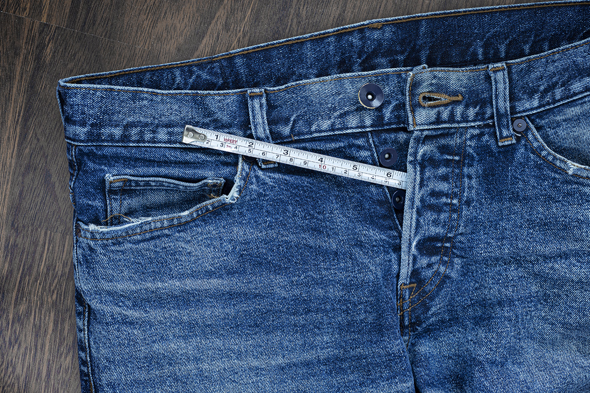 A pair of unbuttoned blue jeans are placed on a wooden floor with a measuring tape poking out of the buttons on the front.