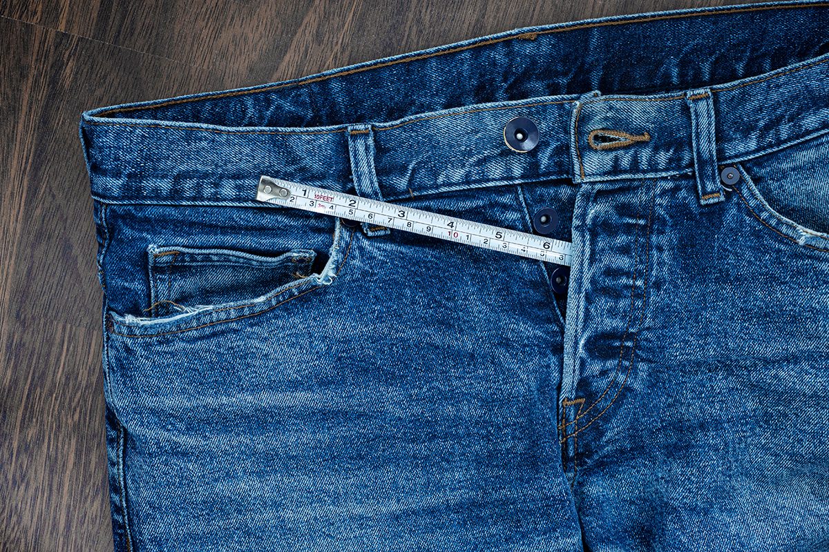 A pair of unbuttoned blue jeans are placed on a wooden floor with a measuring tape poking out of the buttons on the front.