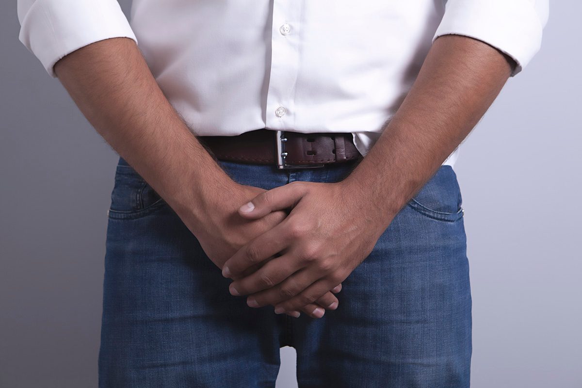 A man wearing blue jeans and a white shirt stands with his hands clasped in front.