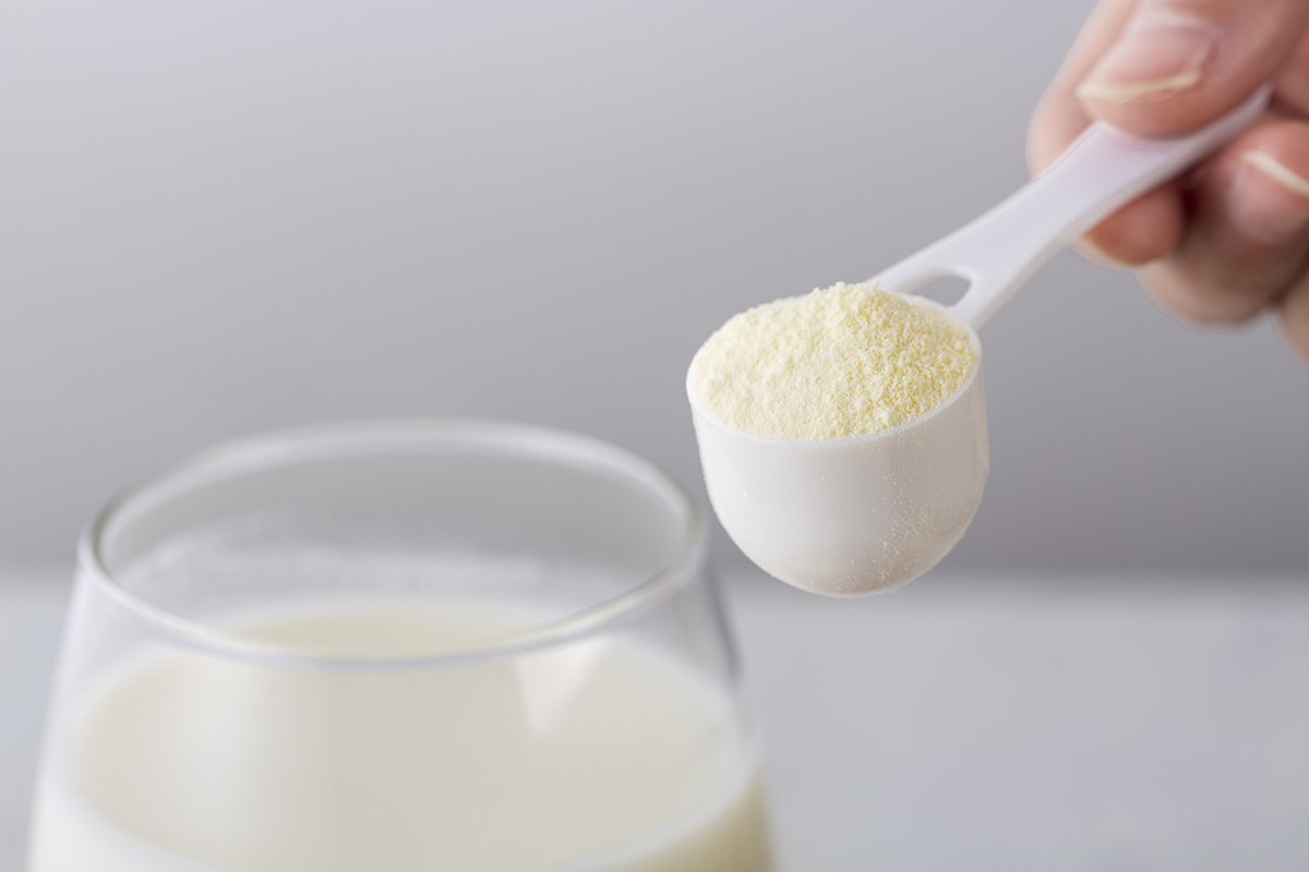 Hand holding a spoonful of powdered supplement next to a glass of milk