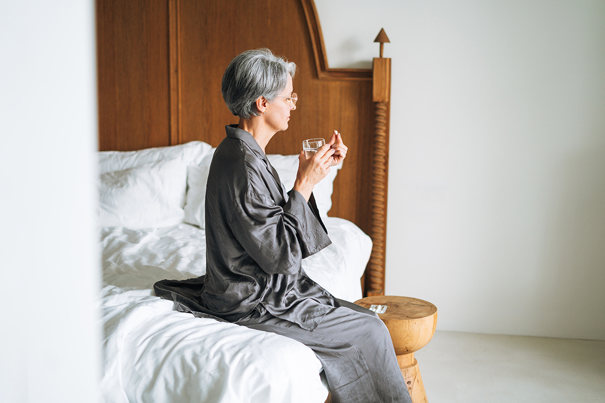 A woman is sitting on a bed holding a vitamin in one hand and a glass of water in the other
