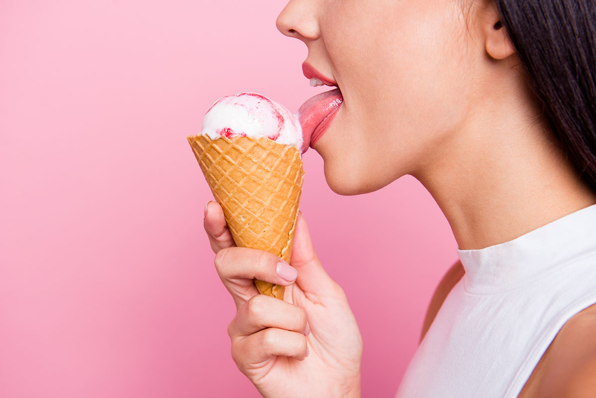The side profile of a woman licking an ice cream against a pink background