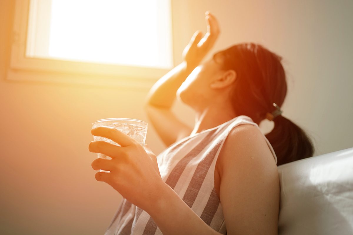 A woman holding a glass of water sits next to a window with light streaming in