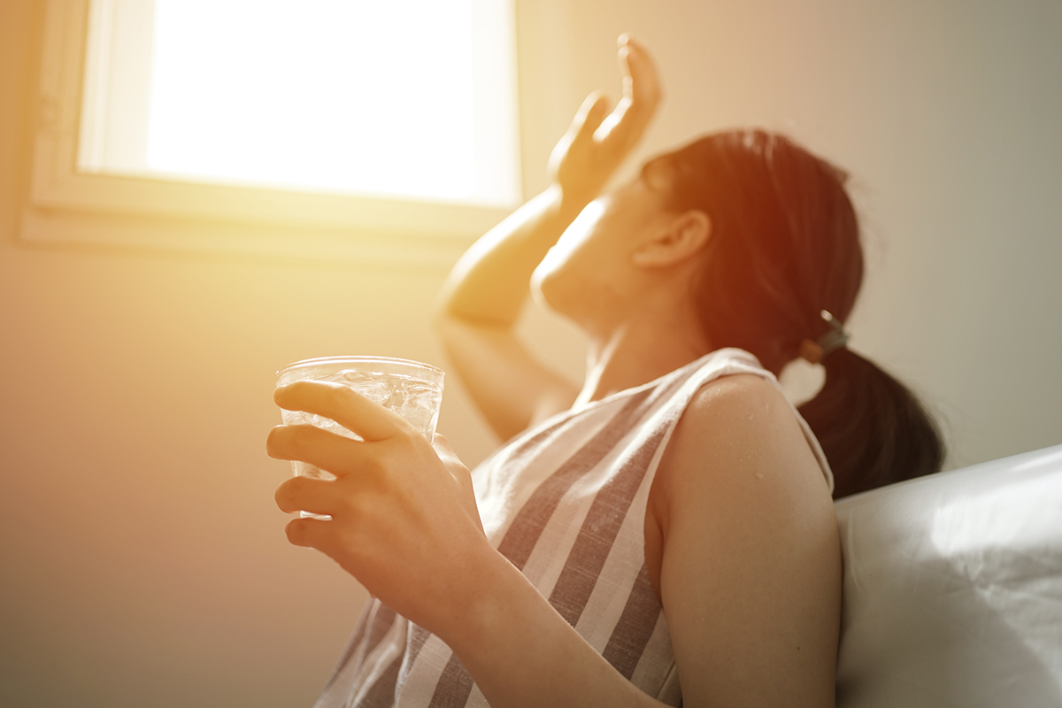 A woman holding a glass of water sits next to a window with light streaming in