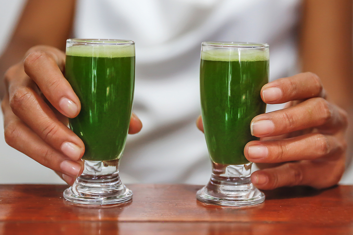 A woman holds two small glasses containing broccoli shots, which are placed on a wooden surface.
