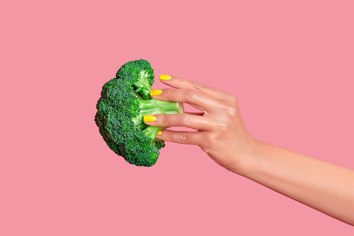 A hand holds a floret of broccoli against a pink background.