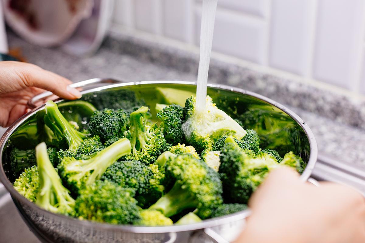 A person is washing chopped up broccoli florets in a metal colander under running water.