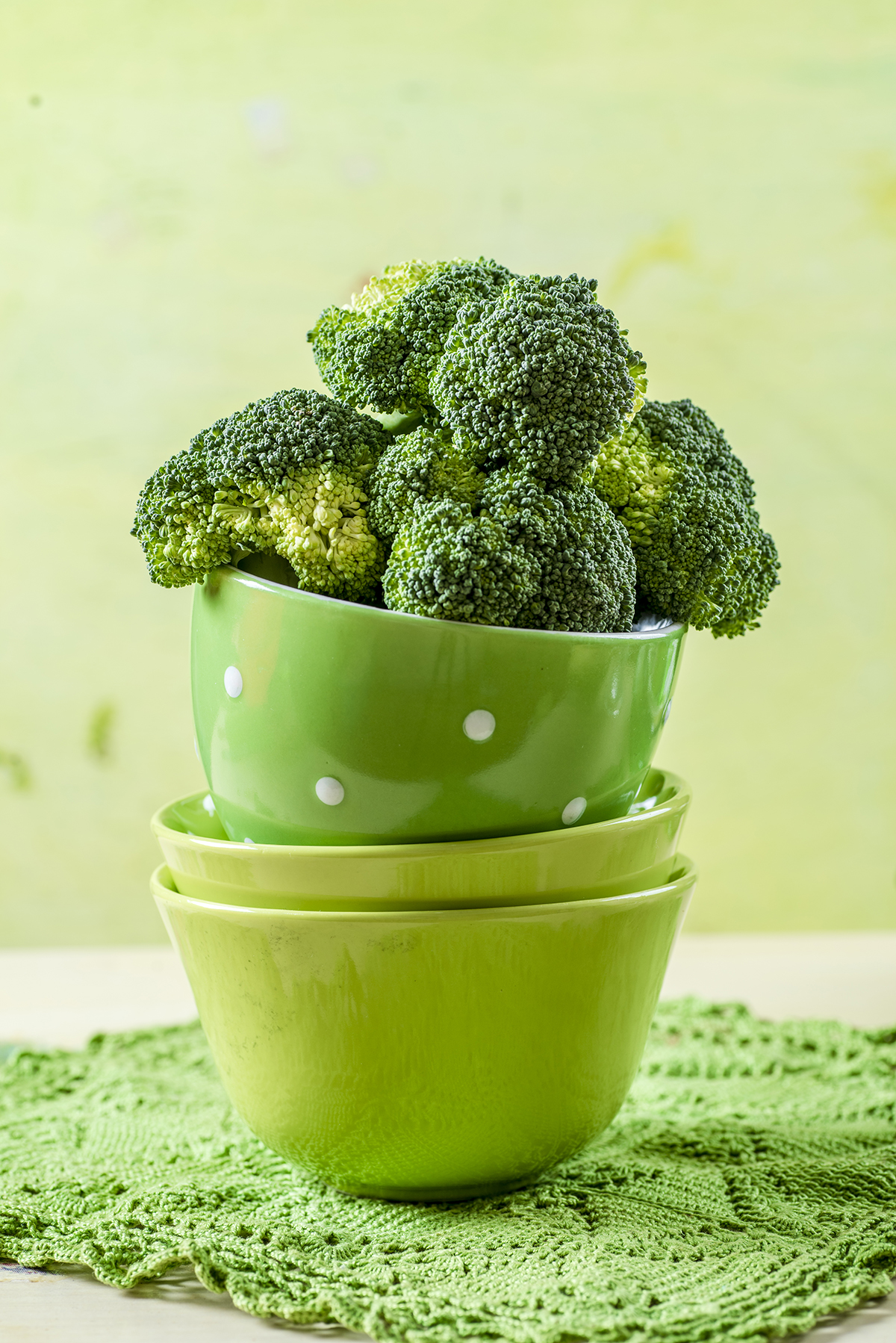 A stack of three green bowls, with broccoli florets filling the top bowl, is set against a green background.