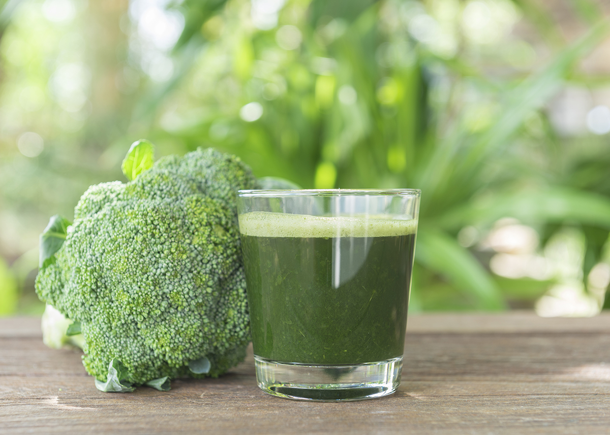 A glass of broccoli juice placed on a wooden table next to a floret of broccoli is shown.