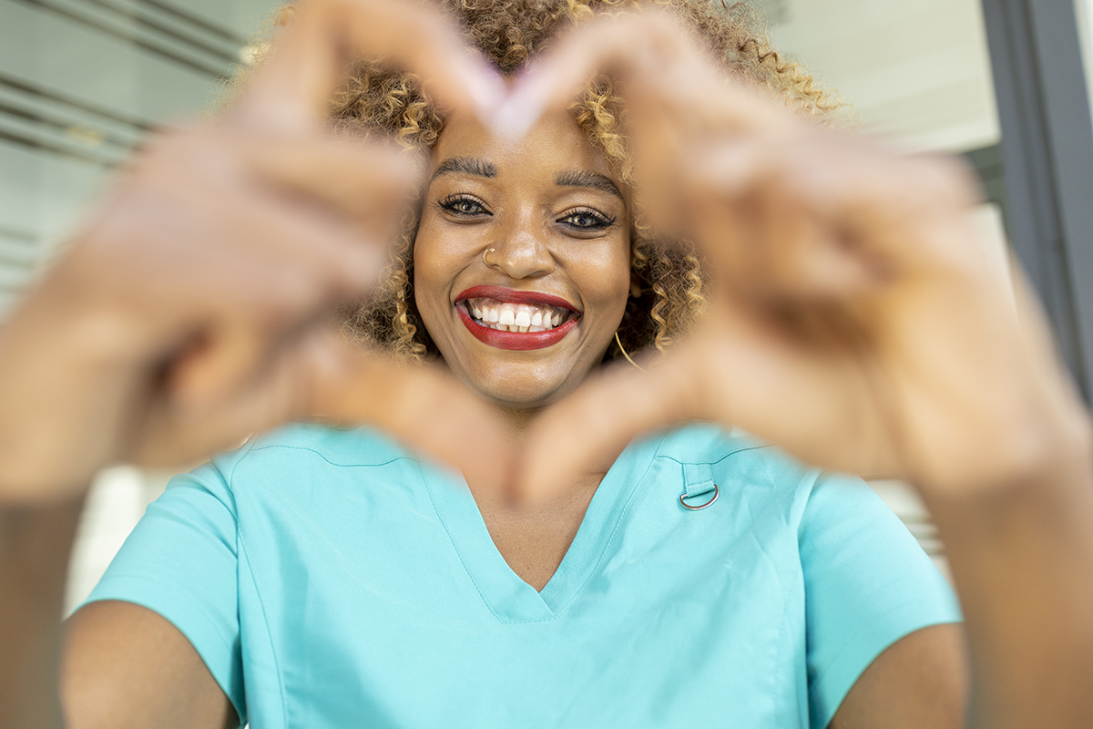 A woman wearing a blue top smiles and makes a heart shape with her hands.