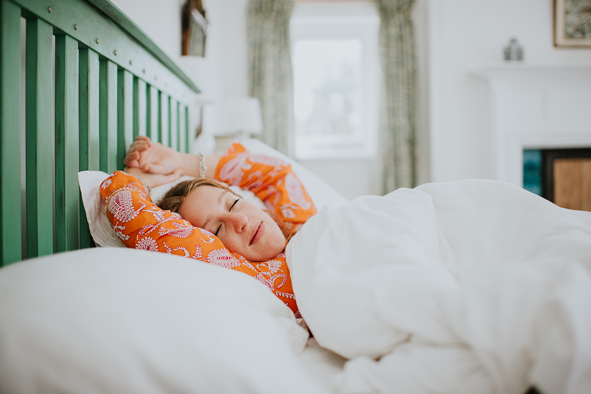 A woman wearing orange pyjamas sleeps with her arms above her head in a bed with white bedding