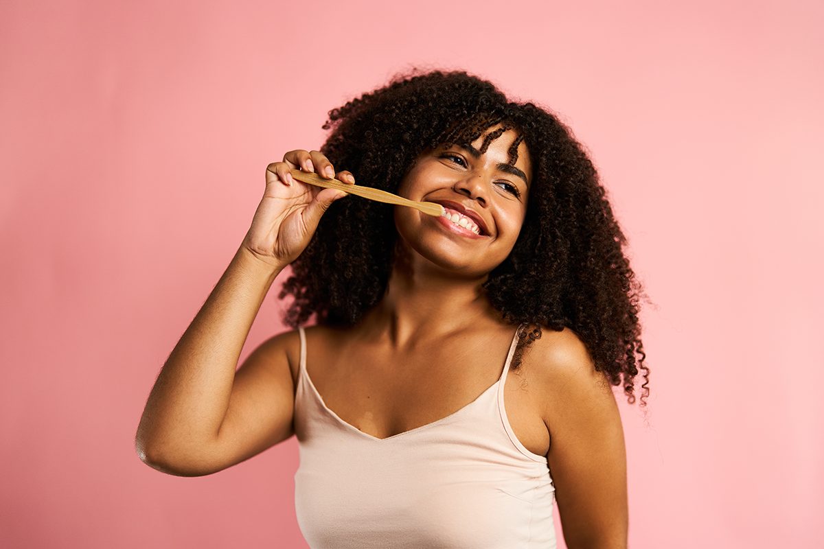 A woman brushing her teeth and smiling against a pink background