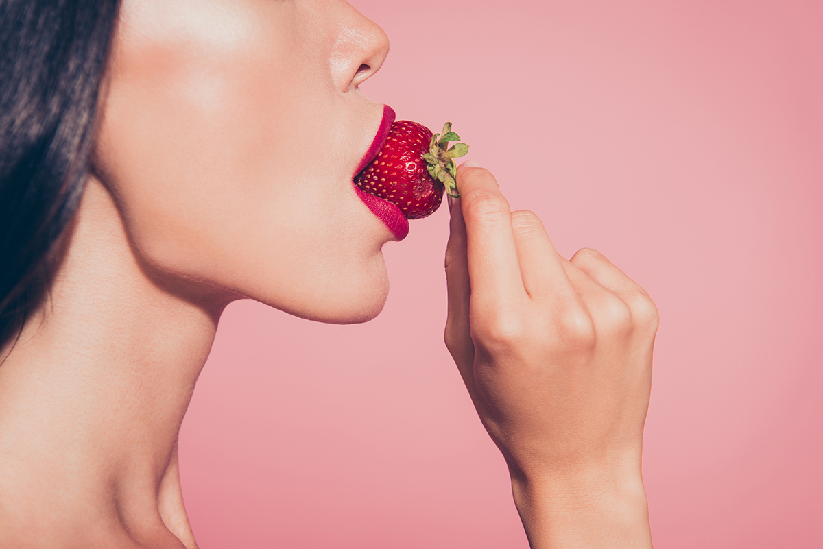 A close-up of a woman eating a strawberry against a pink background