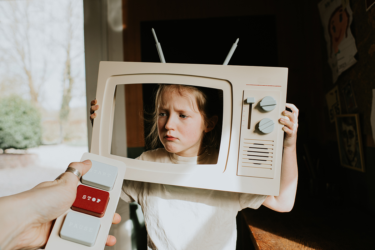 A hand points a remote at a young girl who has a toy television in front of her face
