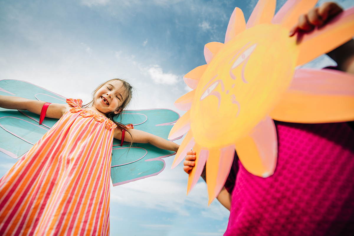 Two children playing outside. One is wearing blue painted wings and the other is holding a cutout sun