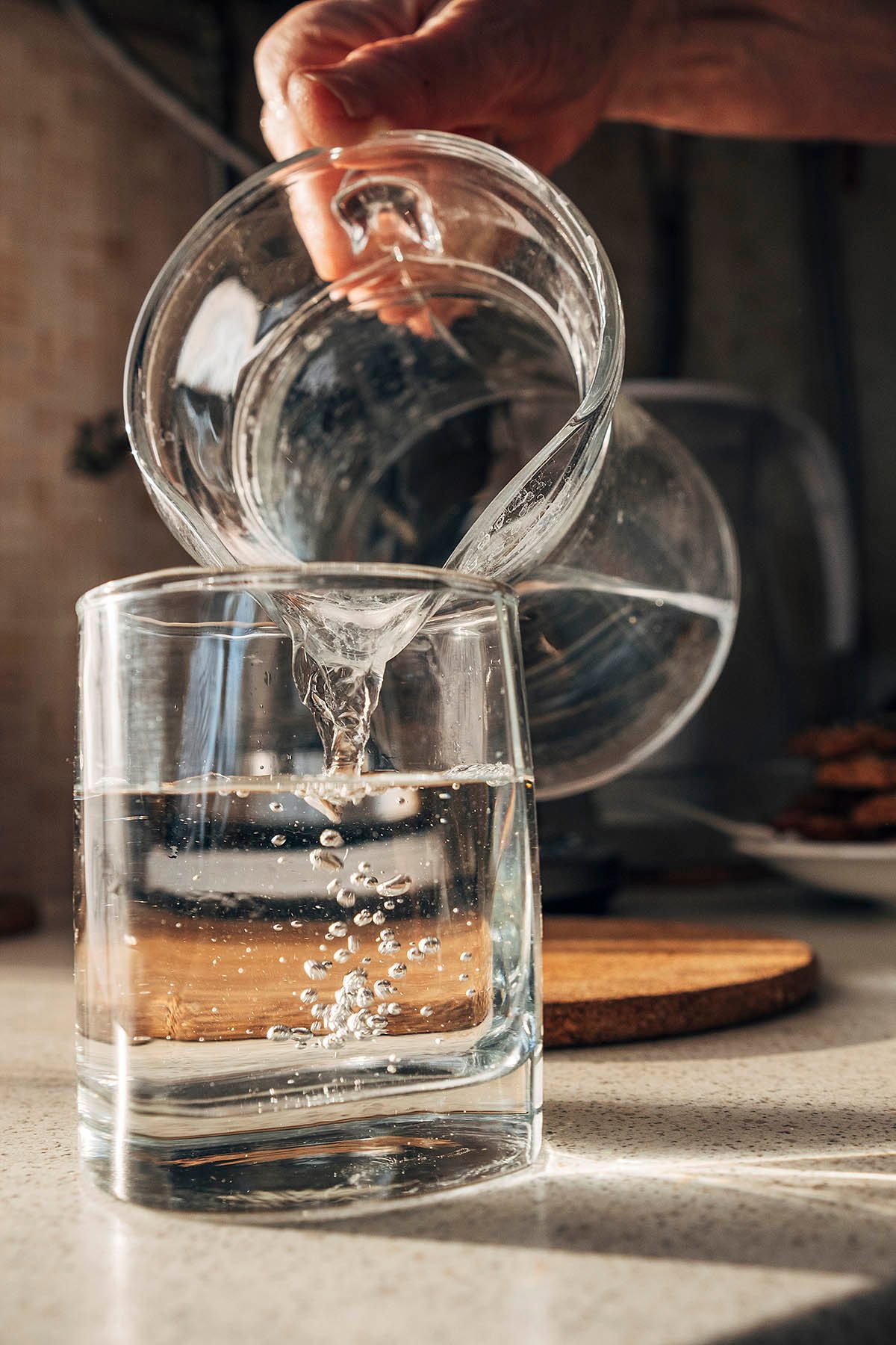 A hand is pouring water from a glass jug into a square glass on a countertop.