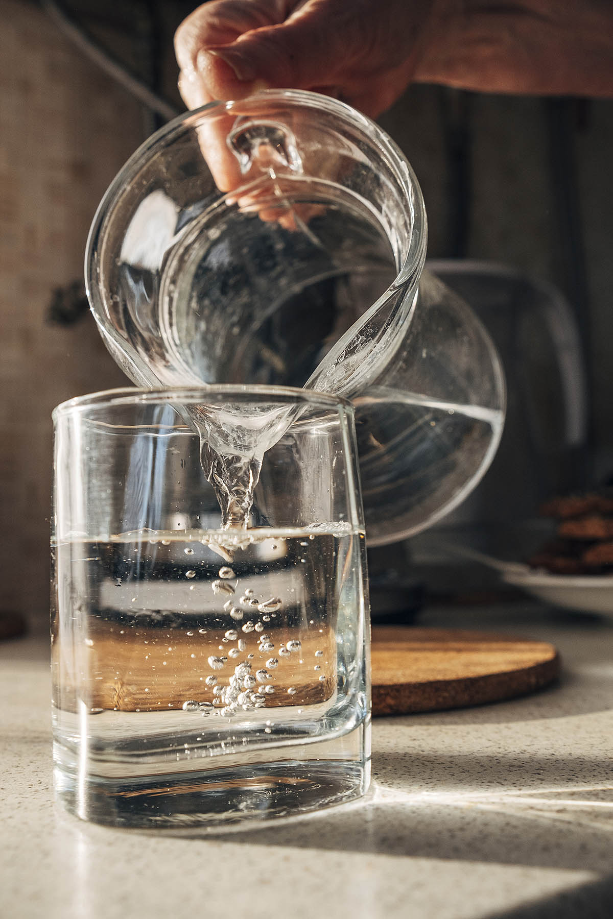 A hand is pouring water from a glass jug into a square glass on a countertop.