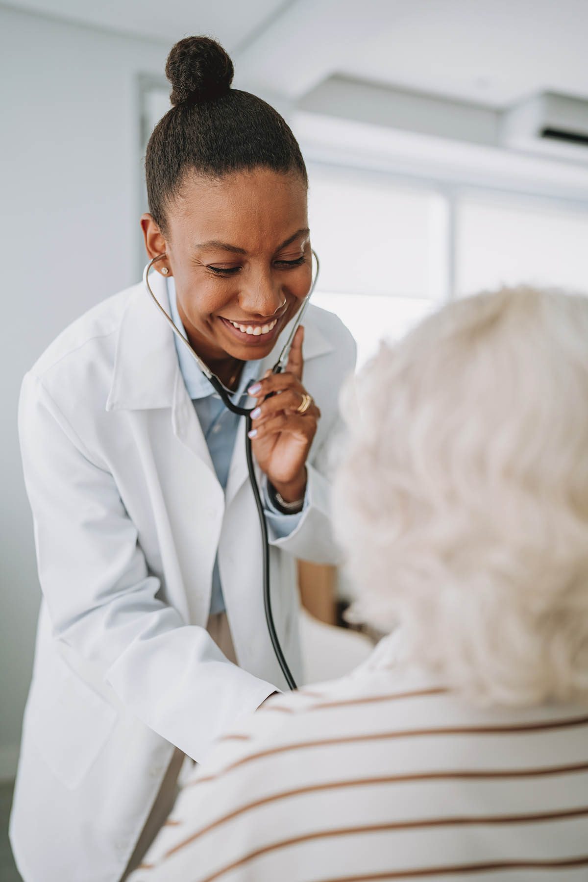 A doctor in a white coat smiles while using a stethoscope to examine an elderly patient.