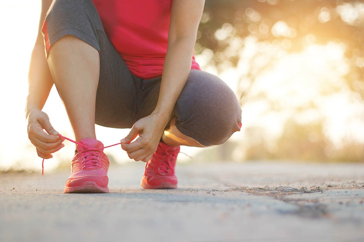 A person in a red shirt and grey shorts crouches down to tie their bright pink running shoes on a street at sunset with a blurred background. s