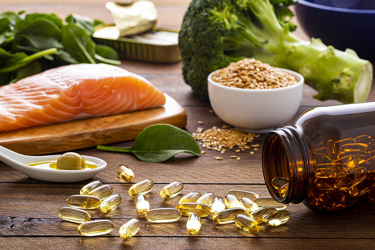 A wooden table displays a variety of healthy foods and supplements, including raw salmon, leafy greens, broccoli, and a bowl of seeds.
