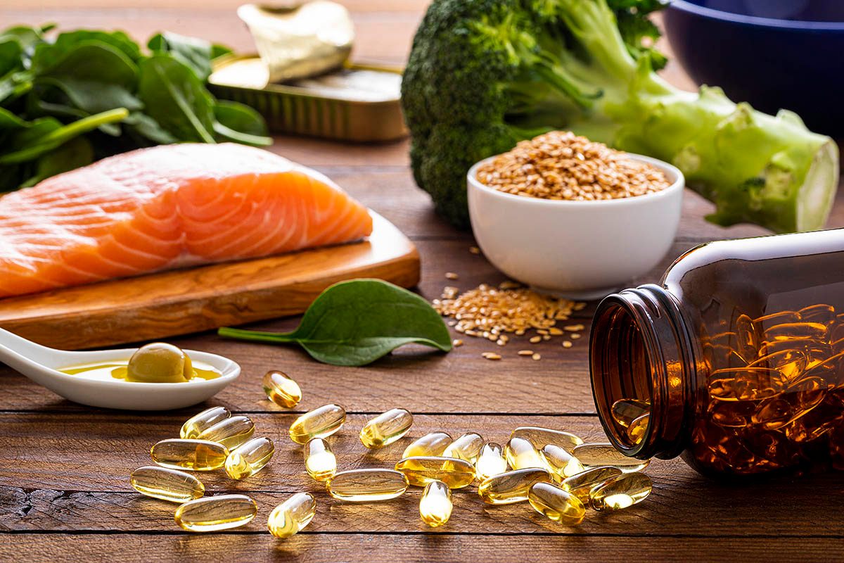 A wooden table displays a variety of healthy foods and supplements, including raw salmon, leafy greens, broccoli, and a bowl of seeds.