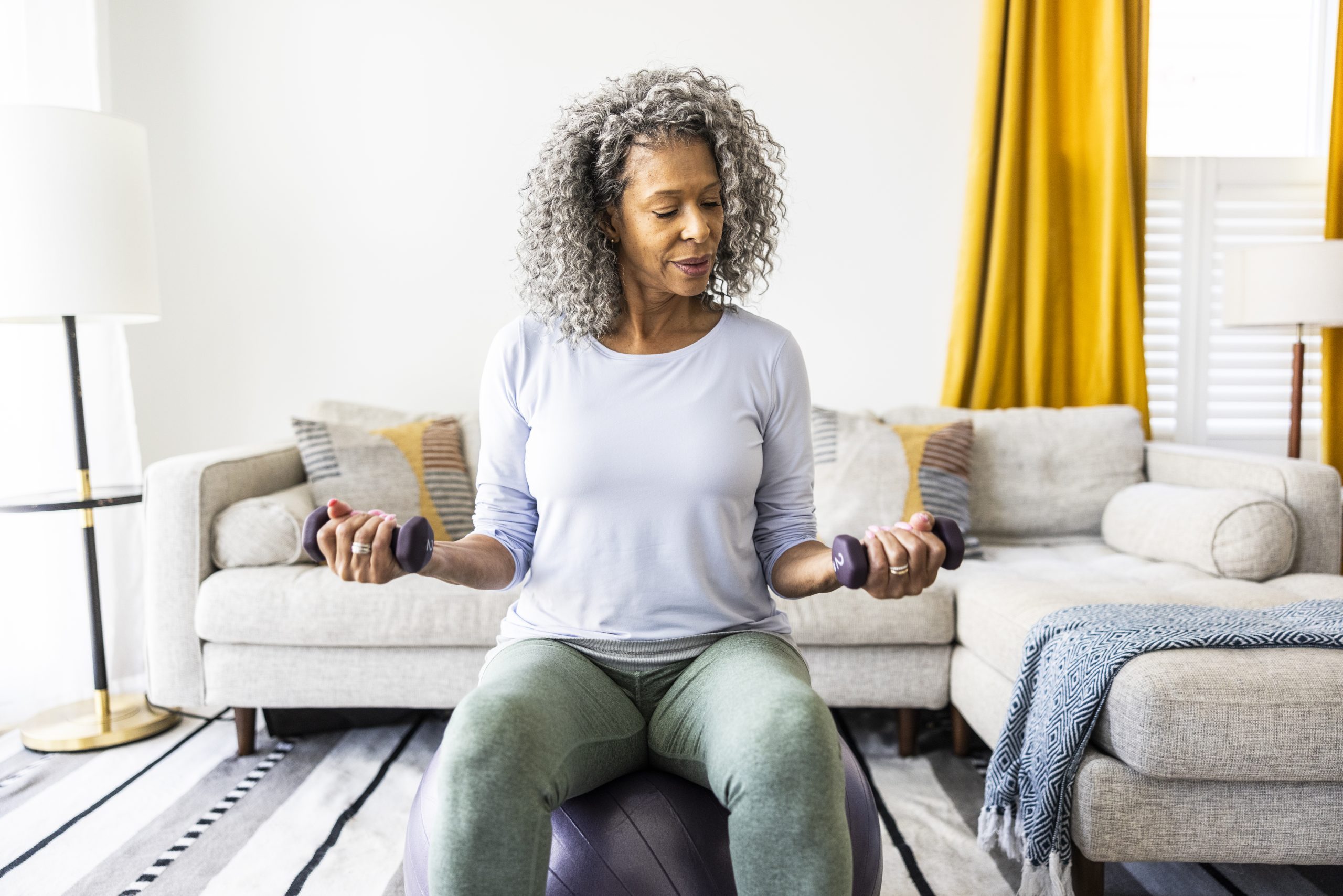 A woman is sitting on an exercise ball, lifting hand weights in a living room