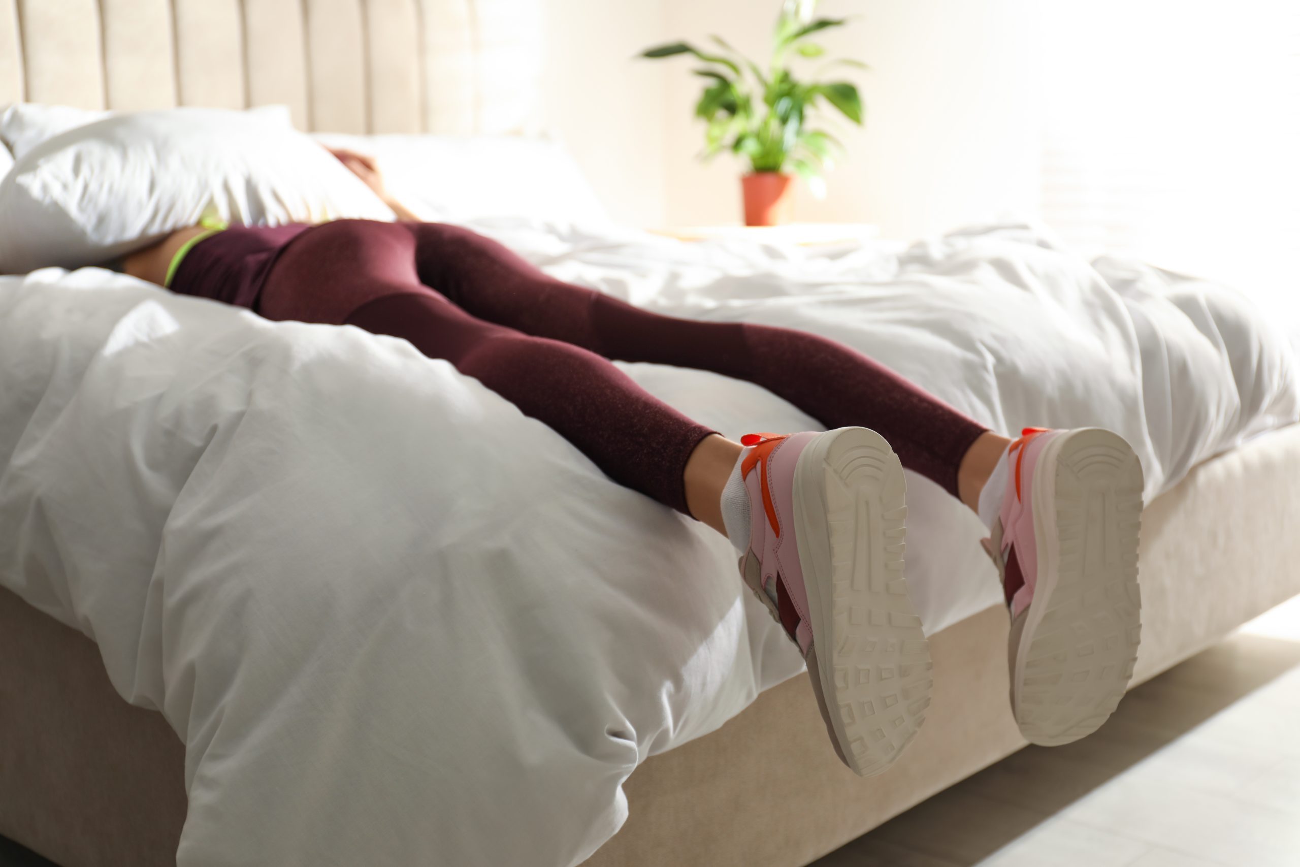 A woman wearing activewear lies face down on a bed, holding a pillow over her head