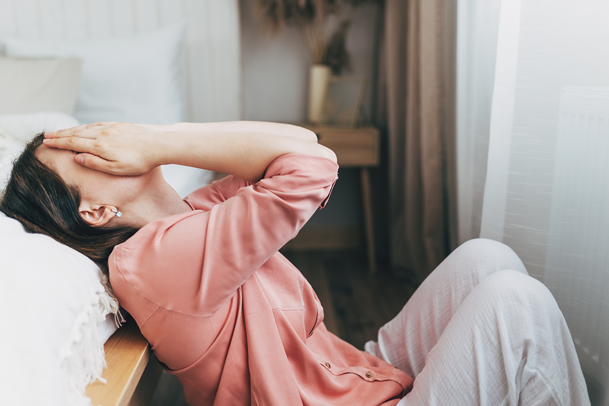 A woman leans against a bed, covering her face with her hands